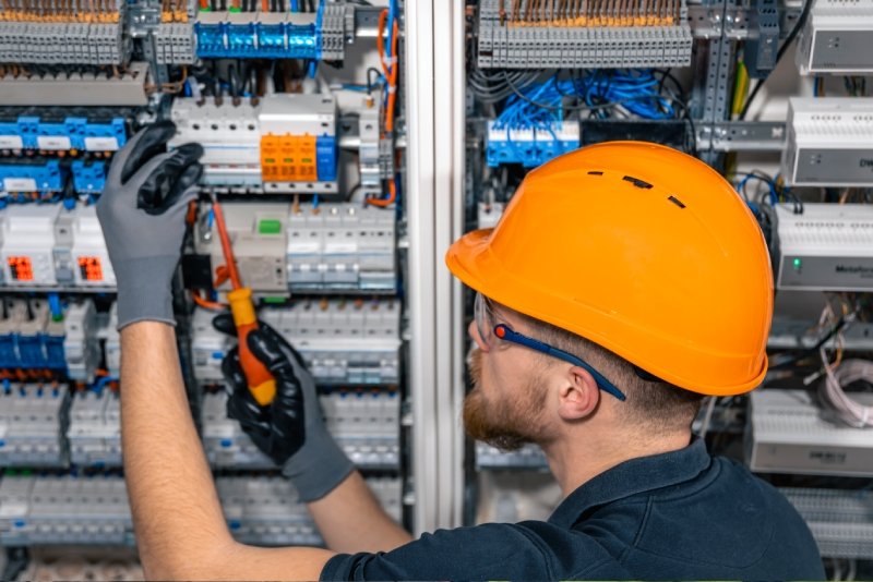 Electrician working on electrical panel with orange safety helmet and gloves, using a screwdriver to repair wiring inside the control panel.